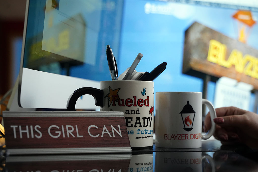 DSC07217 a desk sign that says 'this girl can' in front of a 'fueled and ready' mug filled with pens, a blayzer mug with coffee and a blurry blayzer monitor in the back